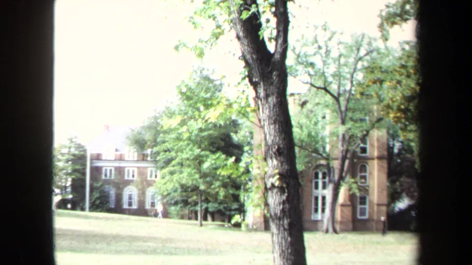 1970 Annapolis campus view with brick buildings, white-trimmed windows, grass, trees, and dark vertical foreground framing.
