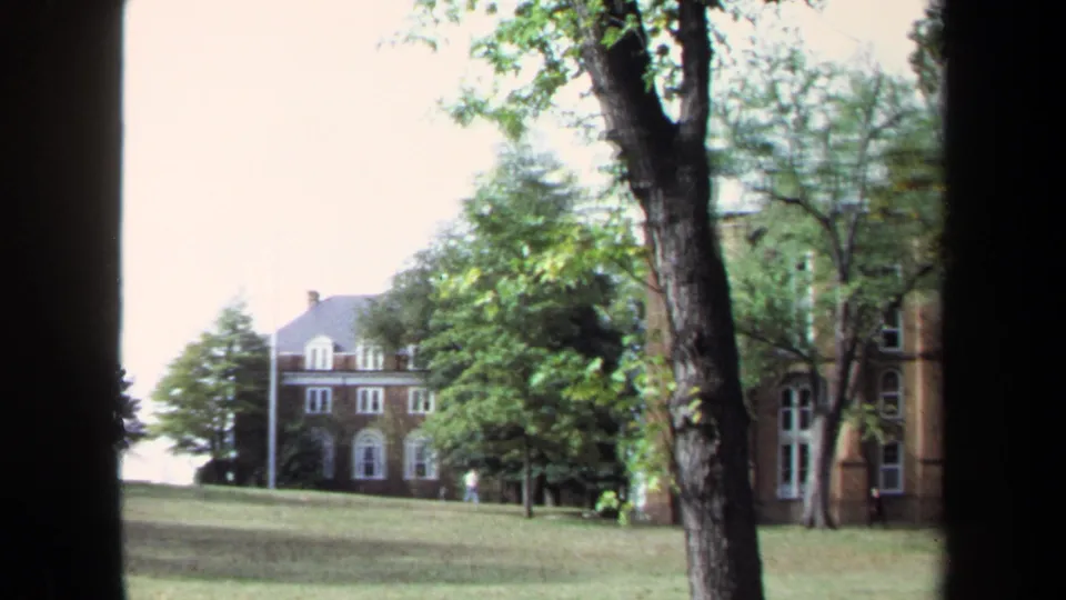 1970 Annapolis campus grounds with a brick building, arched windows, trees, open lawn, and distant pedestrian activity.