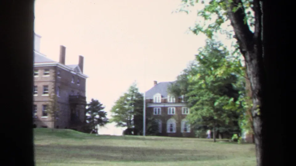 1970 Annapolis campus scene showing brick institutional buildings, a flagpole, trees, lawn, and dark foreground framing.