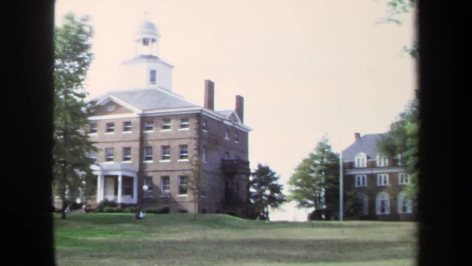 1970 Annapolis campus view of a brick building with cupola, columned portico, chimneys, lawn, trees, and distant people.