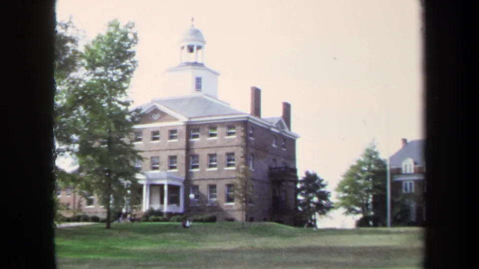 1970 8mm view in Annapolis of a brick institutional building with a white cupola, columns, trees, and lawn, framed by dark film edges.