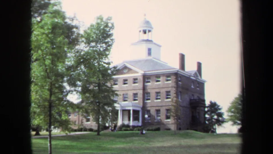 A slightly shifted 1970 home-movie frame shows the Annapolis brick building, white cupola, portico, trees, and open grass.