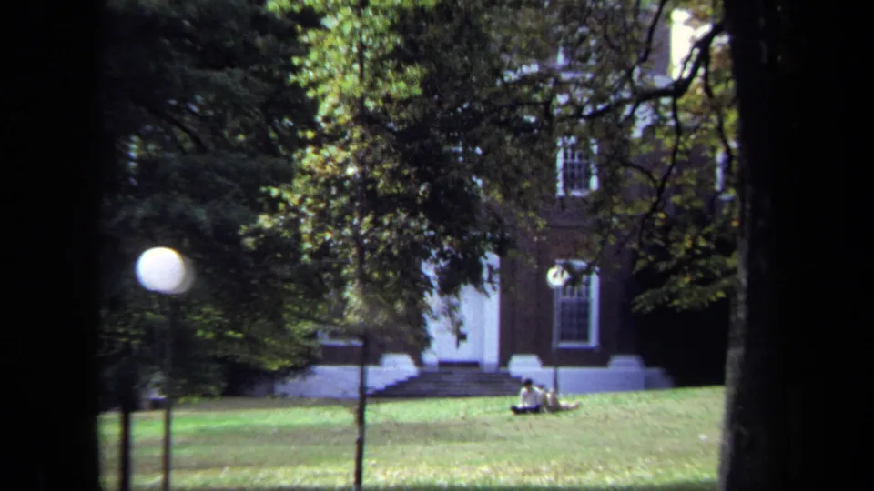 1970 Annapolis home-movie frame showing a brick building with white trim beyond trees, lawn, lamp post, and distant seated people.