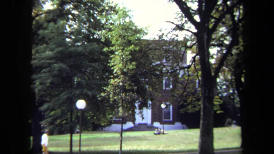 1970 Annapolis frame with trees around a brick building, a globe lamp, seated people on the grass, and a distant walker.