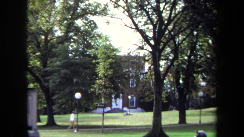 Wider 1970 Annapolis home-movie frame showing a lawn, mature trees, lamp posts, distant seated people, and a walker near the path.