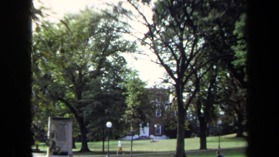Final 1970 Annapolis frame with a broad green lawn, large trees, globe lamp, distant brick building, seated figures, and a walker.