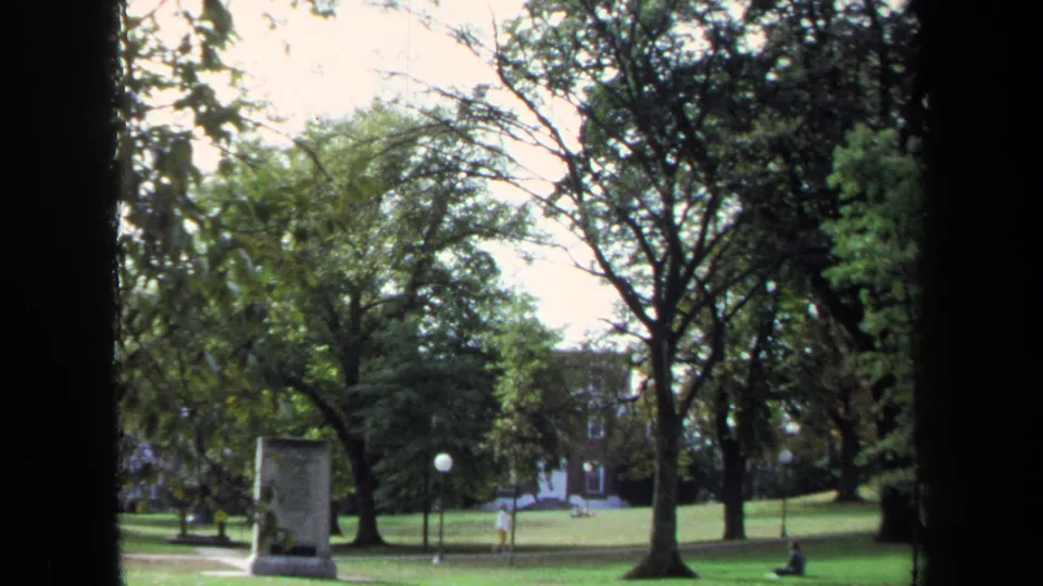 1970 Annapolis 8mm frame showing a green park lawn, mature trees, a stone monument, lampposts, and distant brick buildings.