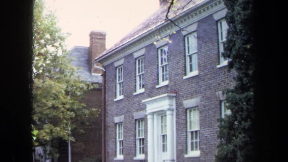 1970 Annapolis home-movie frame showing a brick building with white trim, sash windows and surrounding trees from a partly obscured viewpoint.