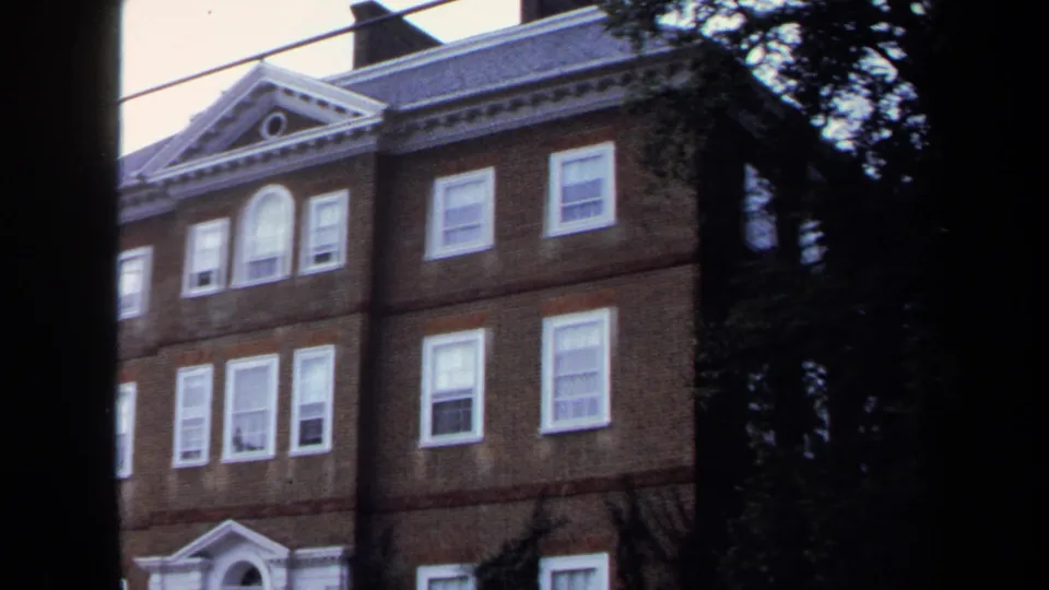 1970 Annapolis 8mm frame showing the upper facade of a brick building with white windows, pediment detail, trees, and dark foreground trunks.