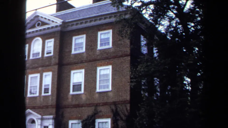 1970 Annapolis view of a brick building's upper stories, with white trim, sash windows, roof pediment, and trees partly obscuring the frame.