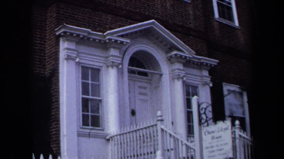 1970 Annapolis frame showing a white portico with columns, an arched doorway, railings, brick wall, adjacent windows, and a small exterior sign.