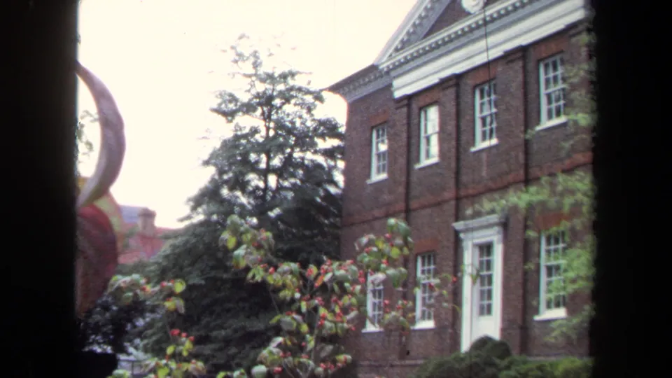 1970 Annapolis 8mm frame showing a historic-looking brick facade, white-trimmed windows, greenery, and dark framing from a nearby window.