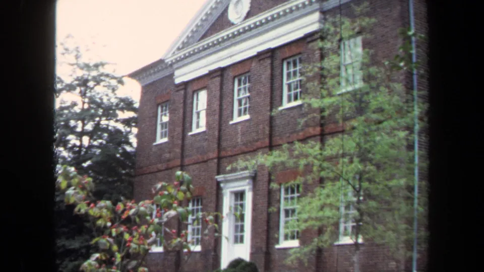 1970 Annapolis 8mm frame with a closer sliding view of white classical trim on a red brick building behind garden foliage.