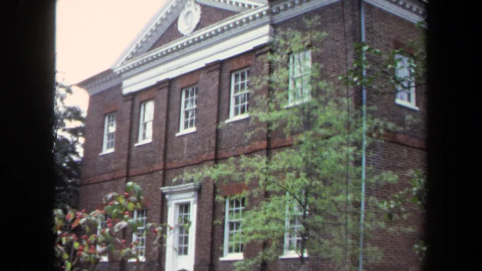 End frame from the 1970 Annapolis clip showing the brick building's white-trimmed entrance and windows through leafy foreground trees.