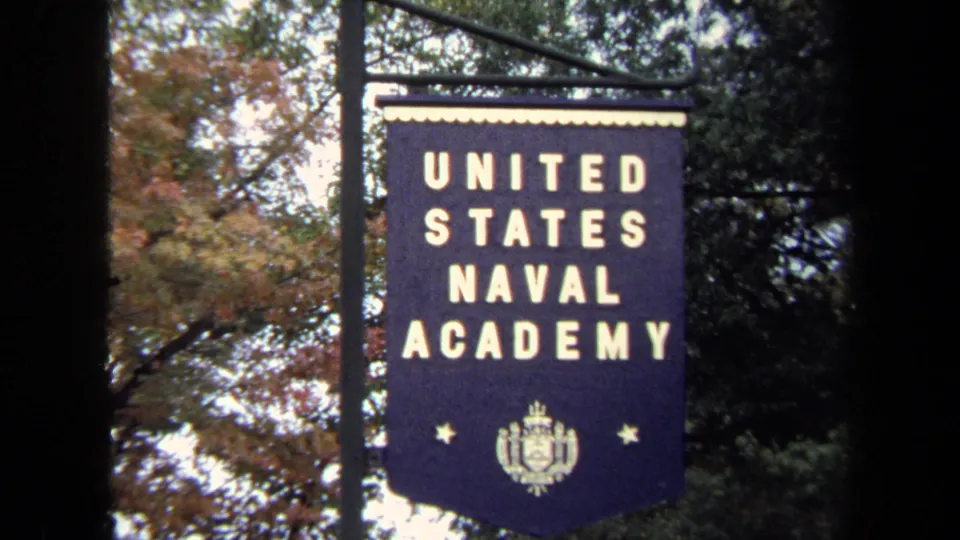 Close visitor-shot view of the United States Naval Academy banner with crest and stars against autumn foliage.