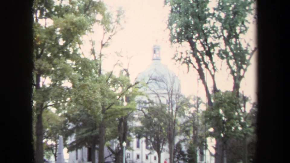 1970 Annapolis frame holding on the white domed Naval Academy building, tree canopy, and overexposed sky in home-movie texture.