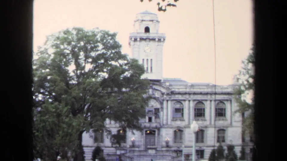 1970 Annapolis home-movie frame of a civic building facade with clock tower, arched windows, trees, and street lamps.