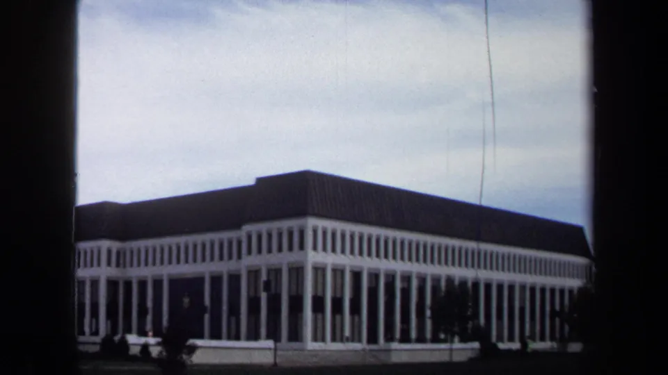 1970 Annapolis home-movie frame showing a large pale institutional building with vertical columns and a dark roof across a lawn.
