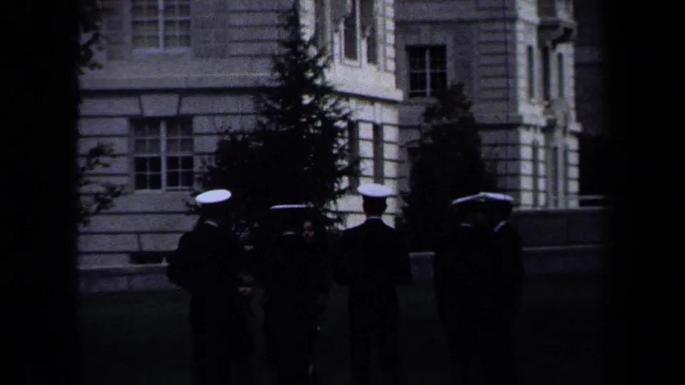 1970 Annapolis home-movie frame with naval officers in dark uniforms and white caps standing near a pale stone building.