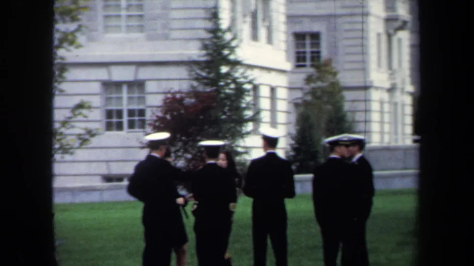1970 Annapolis frame showing several naval officers in white caps standing in conversation on a lawn near a pale building.