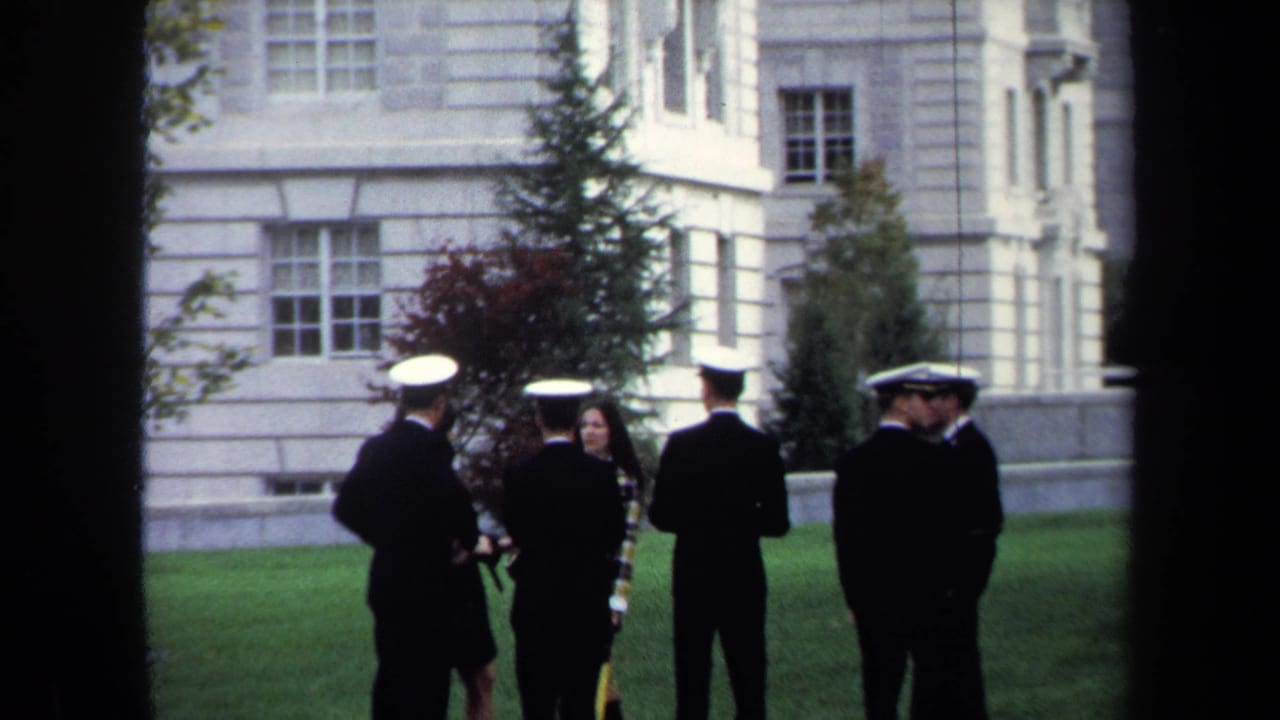 1970 Annapolis home-movie frame of uniformed cadets and a woman talking on a manicured lawn before a pale historic building.