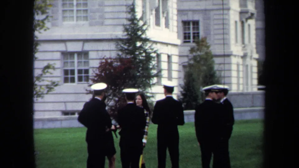 1970 Annapolis home-movie frame of uniformed cadets and a woman talking on a manicured lawn before a pale historic building.