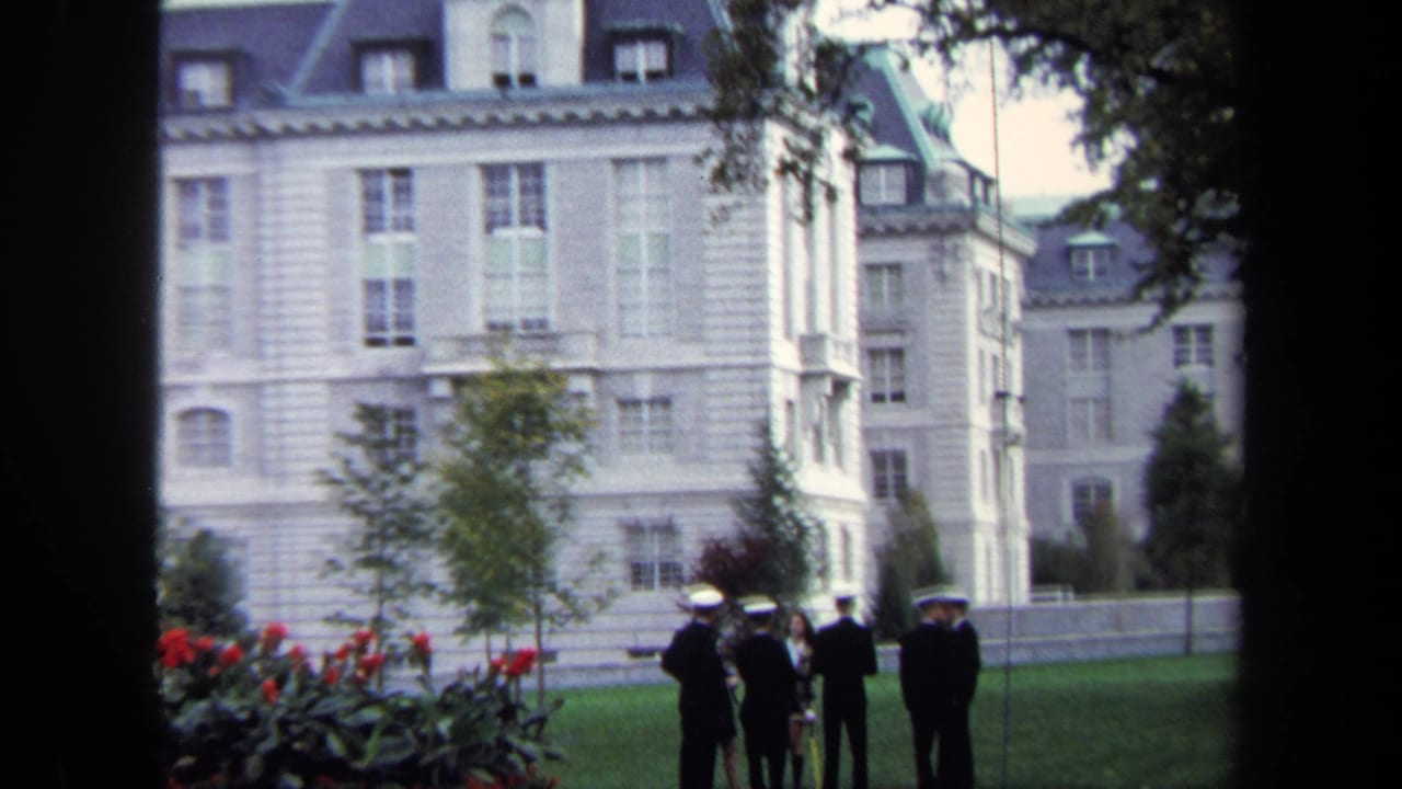 Wide 1970 Annapolis frame showing cadets in white caps gathered near a flowerbed and large historic-looking stone building.