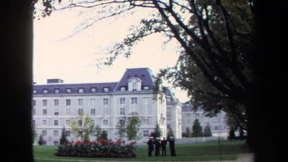 Wide 1970 Annapolis frame of uniformed cadets on landscaped grounds in front of a pale institutional building.