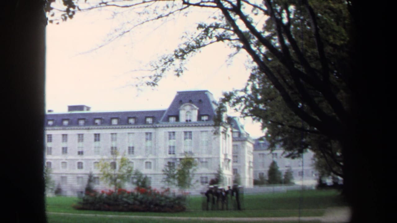Wide view of a pale stone building across a lawn with trees, red flowers, and small distant people.