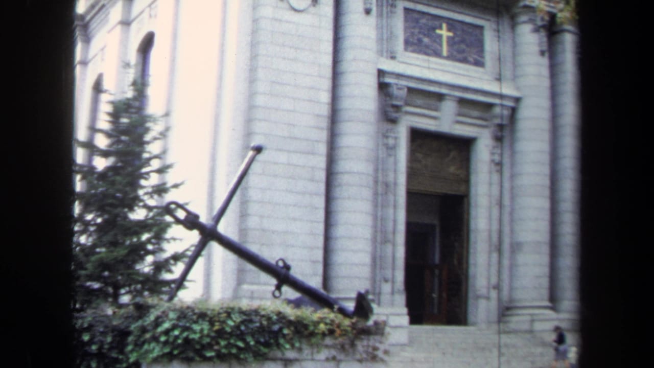 1970s Annapolis 8mm frame showing a white stone church facade, gold cross above the door, trees, and a large anchor monument.