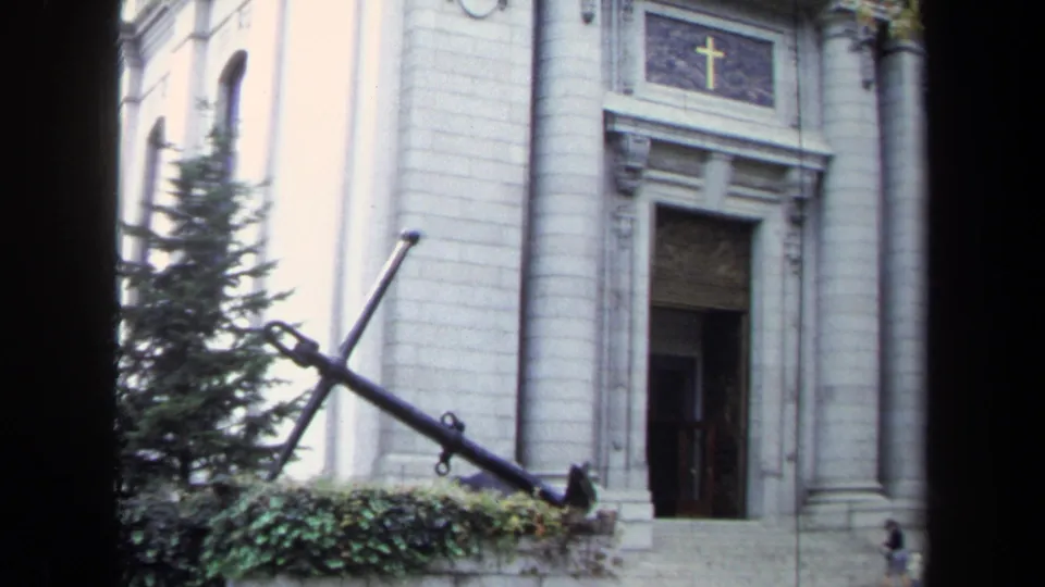1970s Annapolis 8mm frame showing a white stone church facade, gold cross above the door, trees, and a large anchor monument.