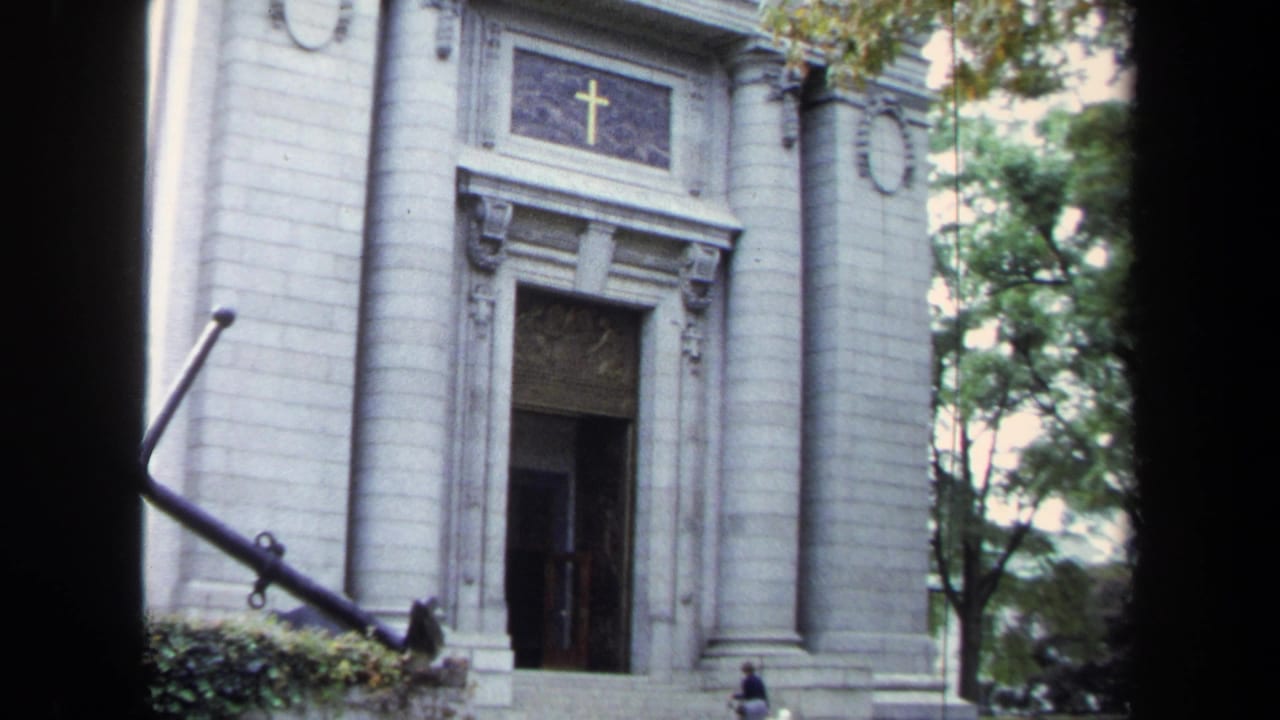 1970s Annapolis home-movie view of the white stone church entrance with anchor monument and small visitors near the facade.
