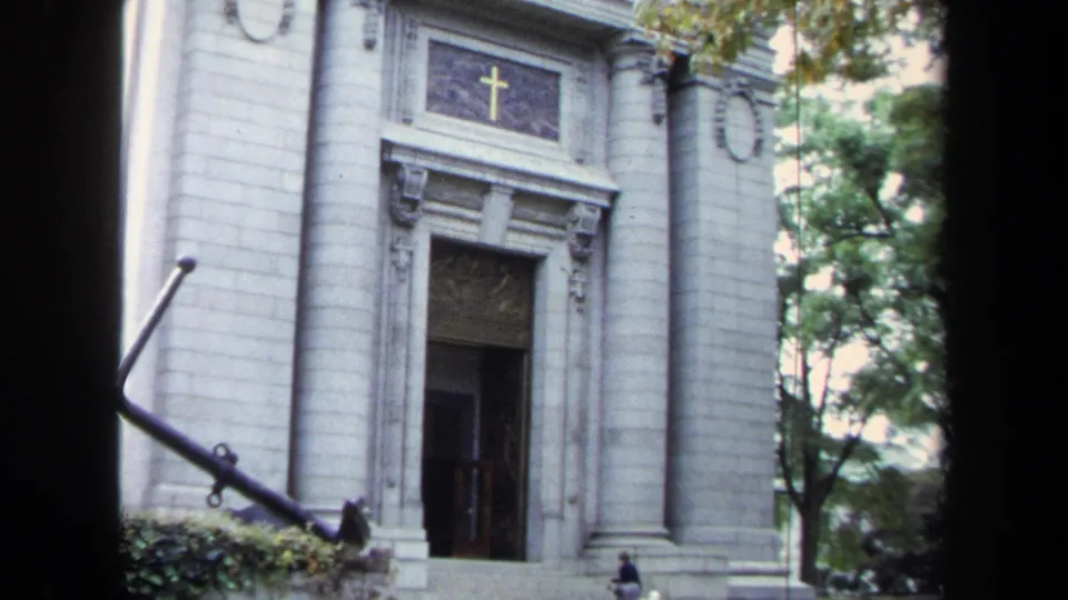 1970s Annapolis home-movie view of the white stone church entrance with anchor monument and small visitors near the facade.
