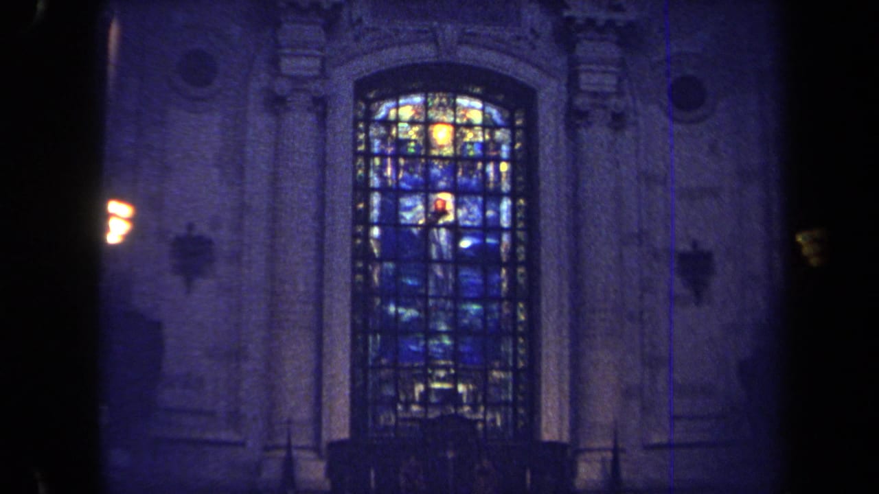 Tall stained-glass window set in carved stonework with wall lights nearby.