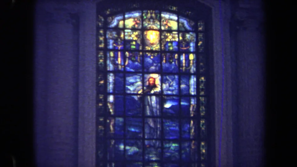 1970 Annapolis 8mm frame of a stained-glass religious figure in blue tones, partly bordered by a dark vertical foreground shape.