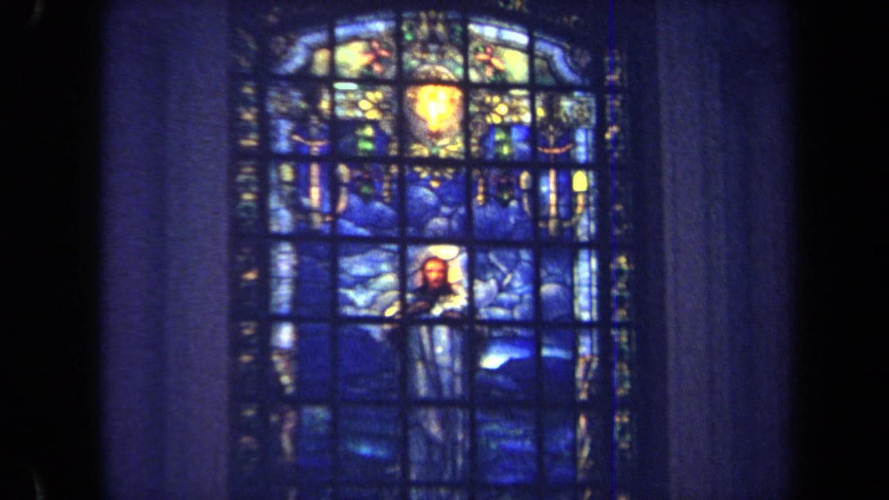 1970 Annapolis home-movie frame showing a blue stained glass church window with a religious figure and heavy interior shadows.