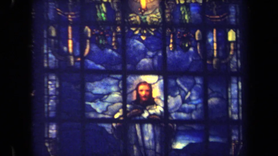 Handheld 1970 church interior view showing the lower portion of a blue stained glass window and a religious figure.