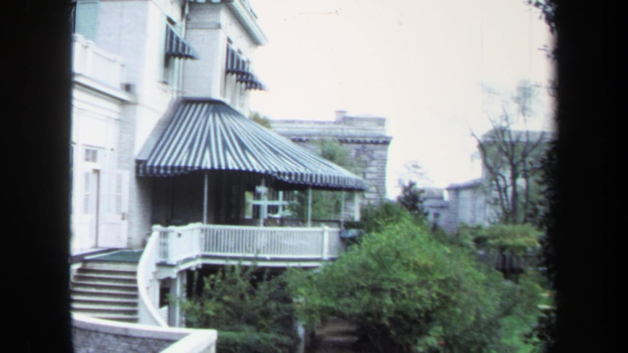 1970 Annapolis home-movie frame showing a large white house with striped awning, porch railings, curved exterior steps, shrubs, and trees.