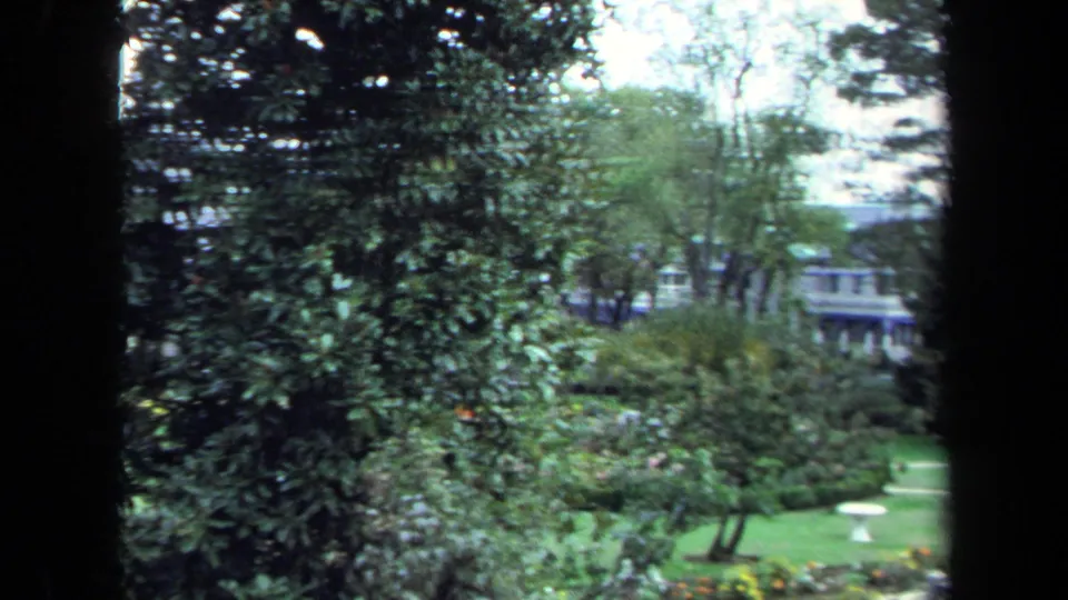 1970 Annapolis home-movie view of a landscaped lawn with trees, shrubs, distant porch structures, and small outdoor tables.