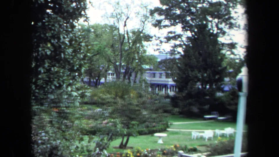 1970 Annapolis frame showing a manicured yard with trees, lawn, white outdoor tables, and distant houses partly behind foliage.
