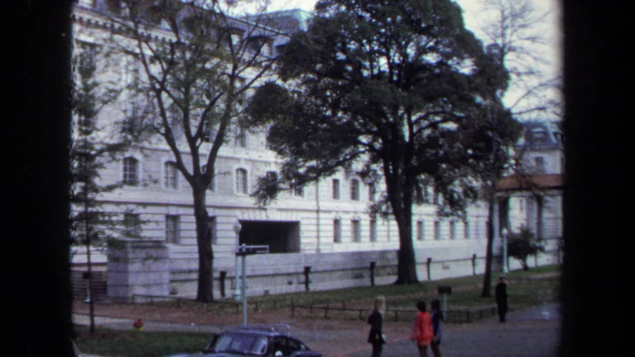 1970 Annapolis home-movie view of a white classical building partly screened by trees, with pedestrians and a parked car near the grounds.