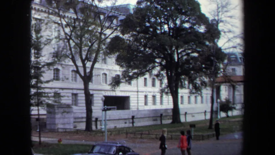 1970 Annapolis home-movie view of a white classical building partly screened by trees, with pedestrians and a parked car near the grounds.
