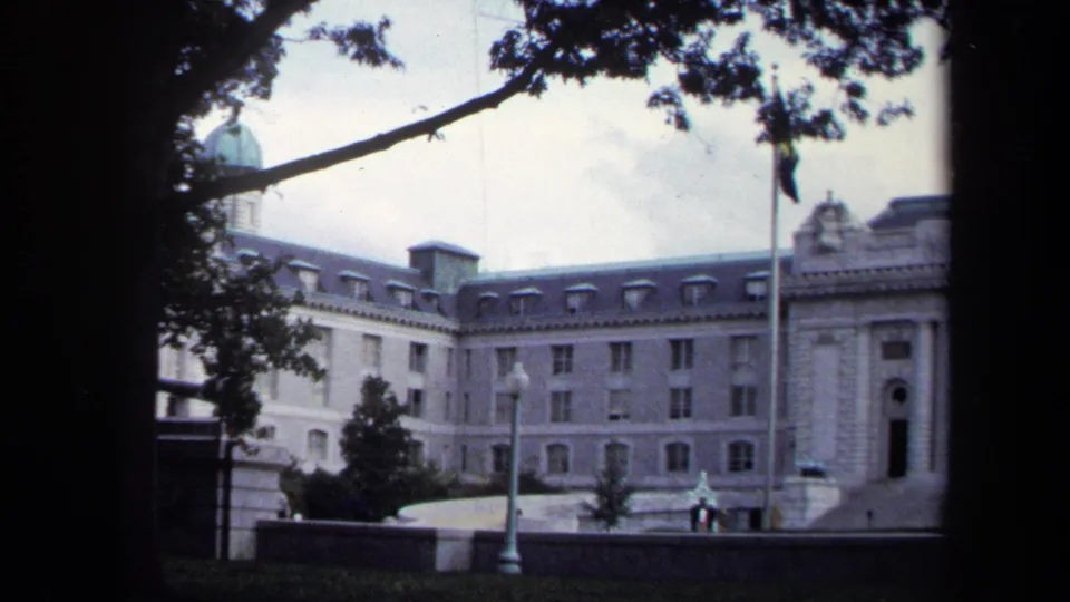 1970 Annapolis home-movie frame of a large white classical building with green dome, flagpole, lamppost, lawn, and distant people near the entrance.