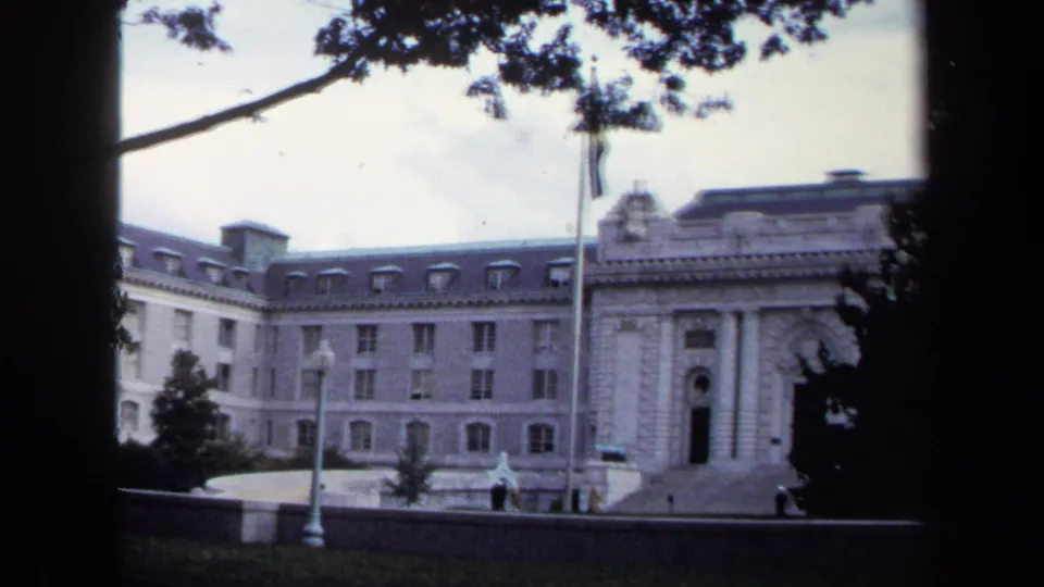 1970 Annapolis footage frames the white classical building, flagpole, lamppost, lawn, tree canopy, and small pedestrians near the entrance.