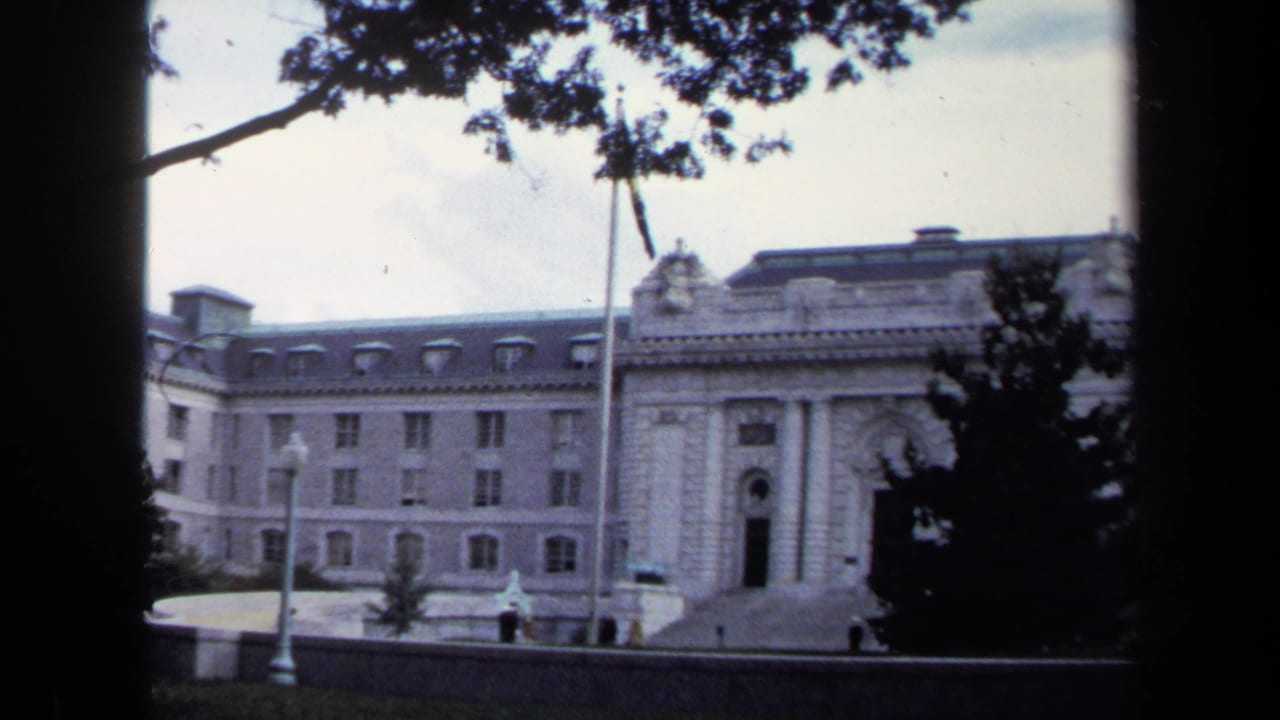 1970 Annapolis home-movie frame showing a formal stone building, flagpole, trees, and overcast sky from a visitor viewpoint.