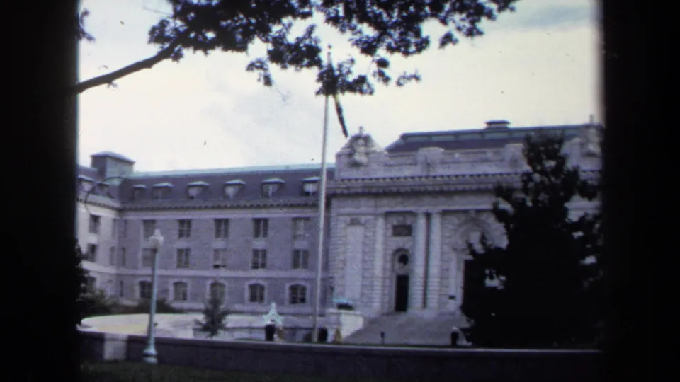 1970 Annapolis home-movie frame showing a formal stone building, flagpole, trees, and overcast sky from a visitor viewpoint.