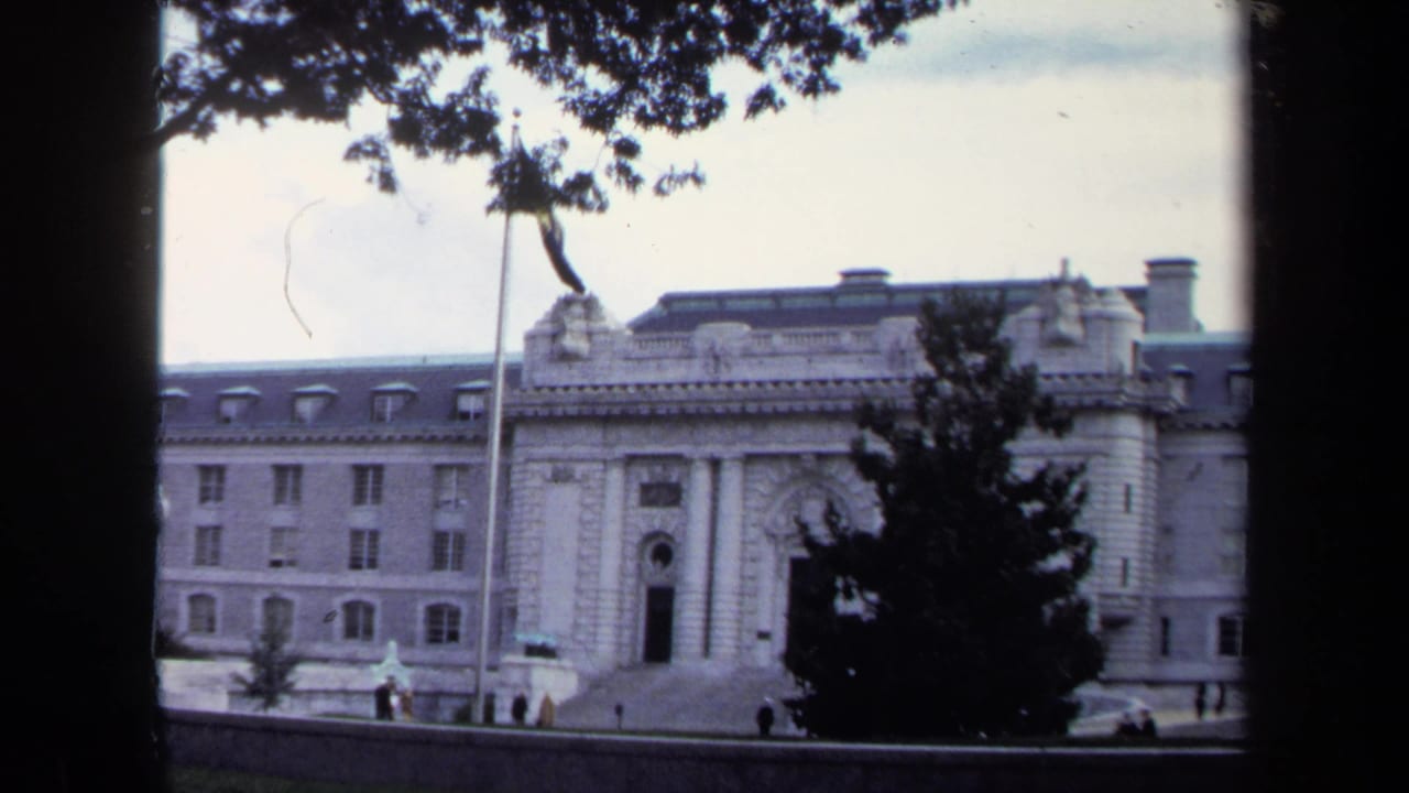 Formal stone facade with flagpole, trees, steps, and small distant figures.