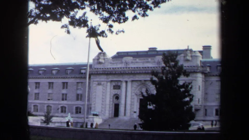 1970 Annapolis frame of a large classical stone building with a flagpole, trees, steps, and distant visitors near the entrance.