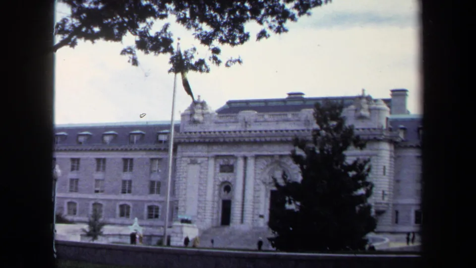 Visitor-shot 1970 Annapolis view of a stone public building with a central entrance, flagpole, tree, walkway, and distant people.
