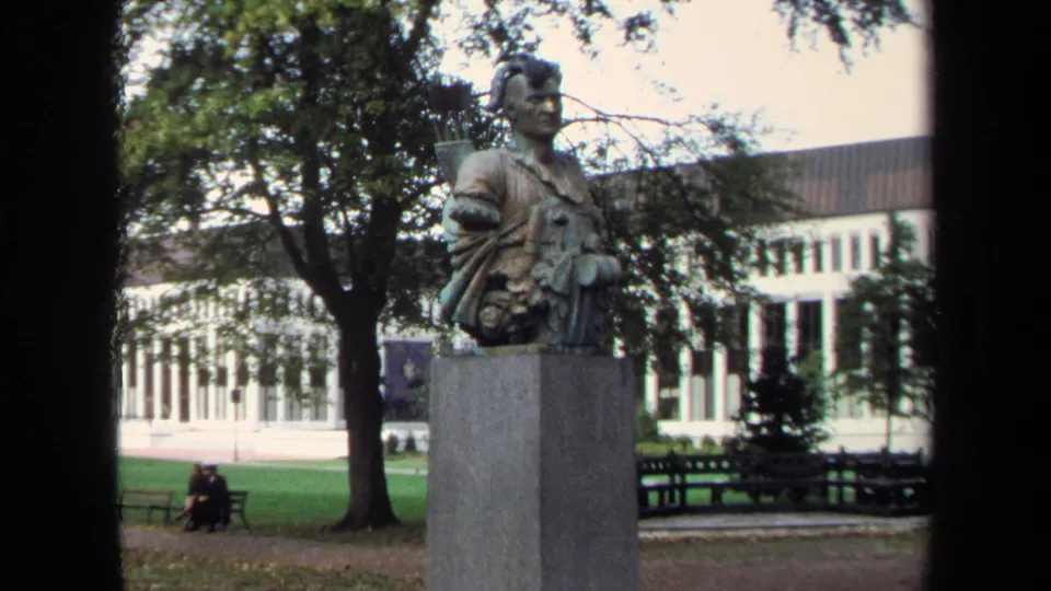1970 Annapolis home-movie frame showing a bronze bust on a stone pedestal in a park-like setting with benches and seated people behind it.